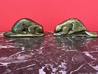 Two brass beaver paperweights shown side by side on marble surface with red background.