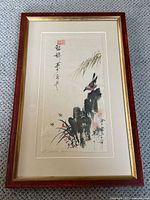 Image showing the entire artwork framed. The watercolor depicts a bird perched on rocks, calligraphy and red seals visible. Frame is reddish wood with gold inner edge.