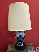 Full view of the lamp showing the white cylindrical lampshade and blue and white porcelain vase base depicting a castle scene. Sitting on a dark ornate wooden base.