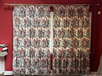 Full view of the floral chintz drapery panels hung on brass rod against red wall, showing the floral pattern, hanging style, and fullness of drapes