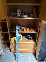 Wooden cabinet with shelves containing various hand tools and hardware including an aluminium bubble level, a large measuring tape, pliers, hammer, and containers with nails and screws.