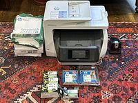 Full front view of HP OfficeJet Pro 8720 printer with multiple new HP ink cartridges in packaging and reams of HP recycled paper beside it, plus electric pencil sharpener on right