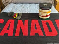 Black towel with red CANADA text, wool hat with pom, and popcorn tin on bed.