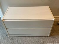 Front view of the white two drawer cabinet showing minimalist flat drawer fronts and white finish, placed on carpet.