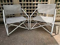 Pair of two vintage Brown Jordan Nomad folding chairs shown side by side outdoors on patio, white aluminum frames and beige fabric seating visible