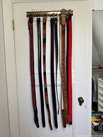 Eight men's belts hanging on wooden rack including leather, woven, and beaded styles in various colors and textures.