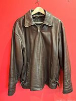 Front view of a men's classic brown leather bomber jacket displayed on a hanger against a red background. Shows full jacket, collar, zippered front, and side pockets with buckle details at the hem.
