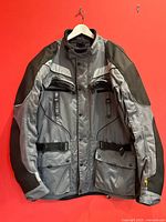 Front view of the men's Olympia AST motorcycle jacket hanging on a hanger, showing grey and black fabric, multiple pockets, and adjustable waist straps.