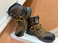 Pair of Merrell men's leather boots, brown and black with yellow laces, showing moderate surface wear. Side angle view of right boot leaning on left boot.