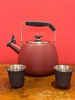 Front view of red Paderno whistling kettle with two black Nespresso double-walled cups in front on wooden table and red background.