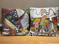 Pair of throw pillows with colorful abstract embroidery and geometric shapes, side by side on a wooden surface.