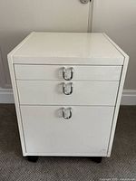 Front view of white 3-drawer cabinet with chrome U-shaped handles and caster wheels.