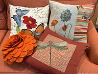 Four decorative throw pillows on a couch including butterfly prints, a dragonfly embroidered pillow, and an orange felt rosette pillow.