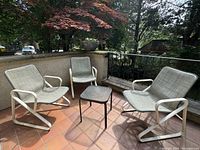Three vintage outdoor sling chairs arranged on a tiled patio with a small table in the middle, showing white metal frames and woven mesh seats.