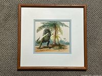 Framed signed painting of a T-Rex dinosaur by Eleanor Kish in a prehistoric setting with palm-like tree, viewed from afar showing full frame and matting.