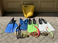 Photo showing four pairs of snorkeling fins in various colors, three snorkeling masks, four snorkels, and one yellow Seal Line dry bag arranged on paved ground near garage door.