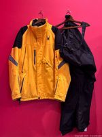 Yellow Spyder ski jacket with black shoulder panels and matching black snow pants hanging on a red background.