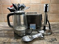 Wide view of lot showing Breville glass kettle, Paderno stainless steel toaster, stainless steel paper towel holder, and large stainless steel spoon in front.