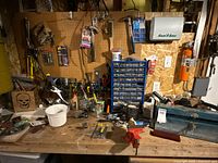 Wide view of workbench with tool clutter including pliers, screwdrivers, hardware bins, and a metal vice mounted on the bench surface.