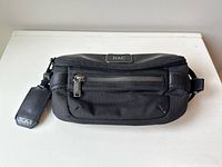 Front view of the black Tumi crossbody bag showing front zippered pocket, DAC patch, and attached Tumi branded tag