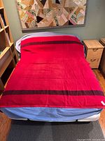 Full view of the red wool blanket spread over a bed showing dark horizontal stripes and overall vibrant red color.