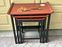 Front view of 3 nested red lacquer tables showing painted Chinese-style scene on top table with sticker residue.