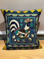Front view of square pillow with colorful embroidered rooster and geometric border on deep blue background.