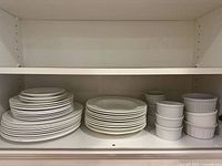 Shelf with stacked white Wedgwood dinner plates, lunch plates, rimmed soup bowls, and white ribbed ramekin dishes in two sizes.