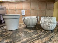 Front view of three white ceramic planters on a kitchen counter with tile backsplash and electrical outlet. The planters include a plain tall pot, a ribbed jardiniere with color grape decoration, and a round planter with floral relief design.