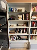 Full view of the white modular bookcase section showing shelves with books, decorative items, and two bottom drawers.