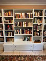 Full frontal view of the white modular bookcase middle section filled with books (not included) showing four shelves above two drawers and two cabinets at bottom.