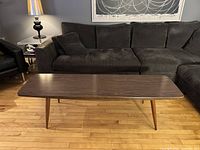 Front view of the mid century modern coffee table in front of a dark sectional sofa. Shows the wood grain laminate top and tapered legs.