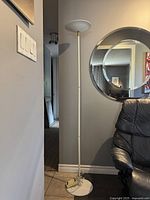 Full view of white metal postmodern floor lamp standing next to a black leather chair, with round mirror on the wall.