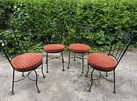 Set of 4 black wrought iron chairs with red round striped cushions arranged outdoors on a concrete surface with greenery in the background.
