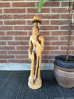 Full front view of the wooden sculpture showing the standing figure with hat and walking stick against a brick wall background.
