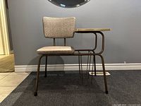 Vintage telephone chair with beige upholstered padded seat and backrest, metal frame, attached wood grain side table and metal wire basket underneath seen from side angle.