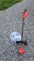 Full view of red cordless trimmer standing on gravel with plastic bag nearby
