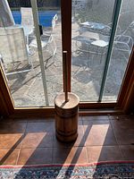 Full view of antique wooden butter churn standing on tiled floor in front of glass sliding door, showing its cylindrical shape and iron bands.