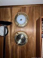 Two brass nautical instruments mounted on wood panel wall, including a barometer on top and a clock below.