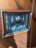 Front view of the wooden framed wall art with nautical knots, pulleys, and a ship model on a blue background.
