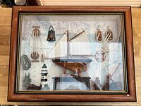 Wood-framed shadow box containing a model sailboat, various ship rope pulleys, coiled ropes, a lighthouse figurine, a life preserver ring, and a lantern, set against a map background.