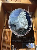 Oval stained glass hanging decoration showing a sailing ship with white sails and blue background.