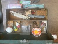 Top view of vintage metal bait box with assorted fishing floats, lures, hooks, weights, and bait placed in compartments