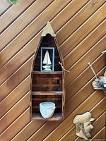 Wooden boat-shaped wall art with two shelves, holding a framed 3D sailboat picture and a ceramic pot.