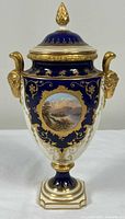 Front view showing cobalt blue and ivory body, gilt decoration, landscape cartouche, ram head handles, lid with finial