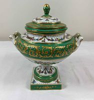 Front view of porcelain covered urn with lid, showing green and gold decoration and floral panels