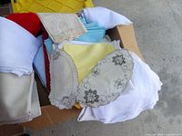 Box of assorted coloured linens including yellow, blue, white cloths and embroidered doilies