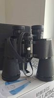 Black textured binoculars with attached neck strap on a box with carrying case behind.