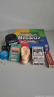Photo showing a collection of assorted games and toys including Blokus board game, ear-shaped model, harmonica, card decks, timer, and tape measure.