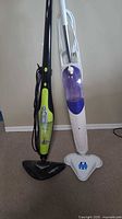 Two upright steam mops standing side by side on carpeted floor, one black and green, the other white and blue with purple tank.